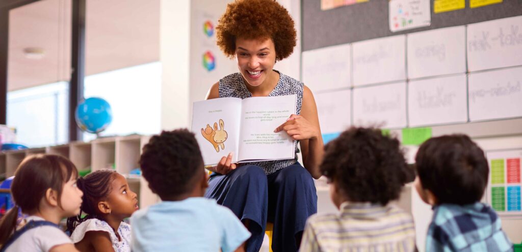 teacher seating in front of a circle of students reading a book